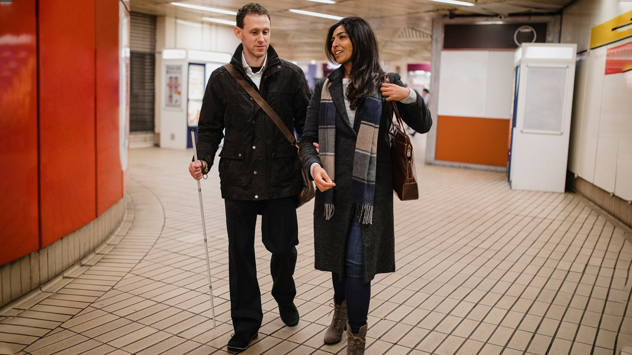 A man with a vision impairment walking with his female friend leaving a subway station, he is using a white cane and is holding his friend's arm for guidance