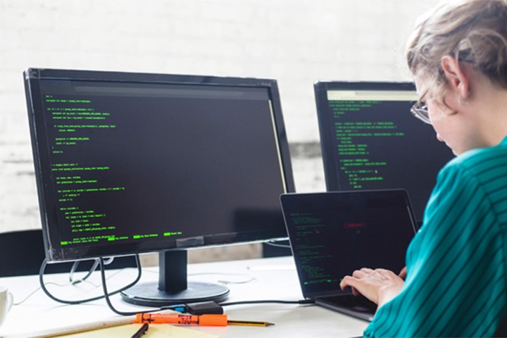 Woman typing computer code with a laptop and desktop computer