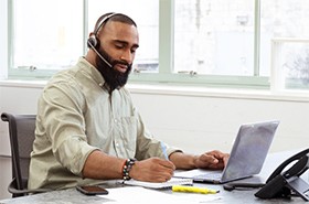Man talking on a headset while taking notes in front of a laptop