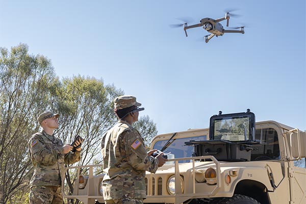Two soldiers in front of a Humvee, one of them is remote control piloting a drone that's flying overhead