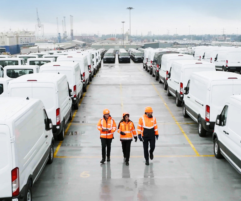 Workers in orange vests and hardhats walk among rows of parked vehicles.