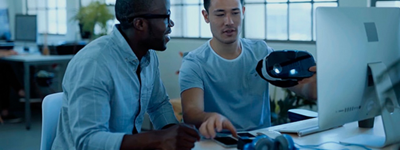 two men talking with a VR head gear in front of PC