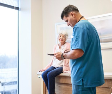 male medical worker holding a mobile device, chart in a room with an elderly woman who is sitting on the exam table