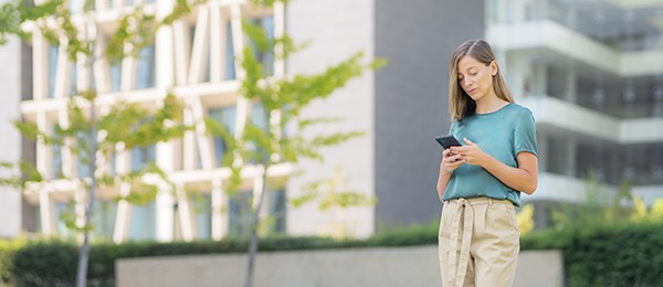 A woman using a smartphone stands outside among tall buildings.