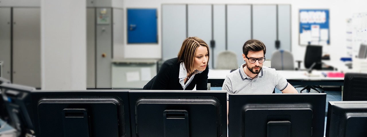 Two coworkers collaborate at a desk in front of a row of computer monitors