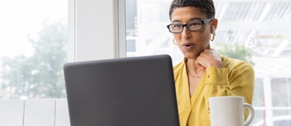 woman wearing a yellow top on a conference call looking at a laptop with ear buds and a coffee cup near by