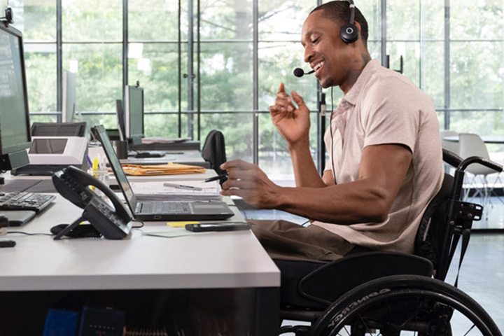Man in wheelchair wearing headsets talking on phone using laptop