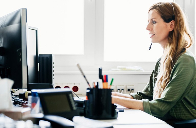 woman sitting in front of two desktop computer monitors wearing a phone headset and typing on keyboard