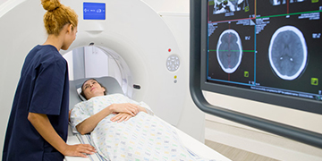 A patient in a CT speaking to the radiology technician with CT scans on the screen.