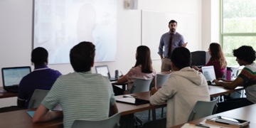 Students in a classroom sit in front of laptops while looking at a projection screen showing a feed of a remote student.